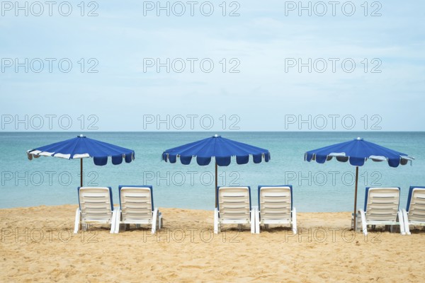 A serene beach scene with three blue umbrellas and white lounge chairs on golden sand facing a