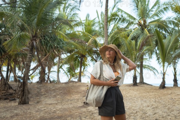 A woman in a summer outfit and hat explores a sandy beach in Thailand, holding her phone under lush