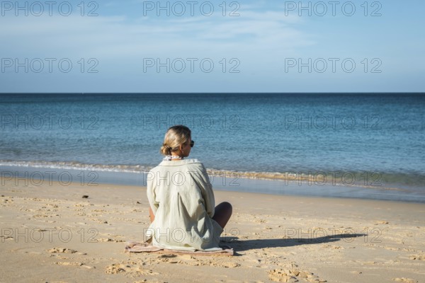 A woman sits calmly on a sandy beach, gazing at the serene ocean under a clear sky in Thailand. Her
