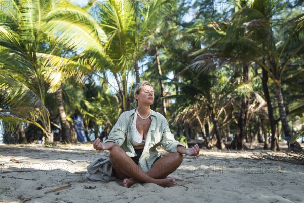 A woman sits cross legged on a sandy beach, meditating peacefully with eyes closed in Thailand.