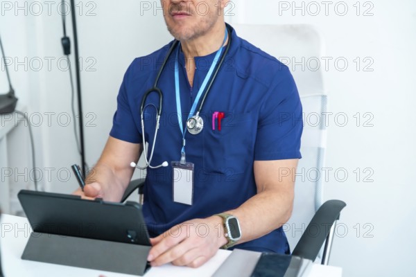 Medical professional in blue scrubs wearing a stethoscope and id card, using a digital tablet while