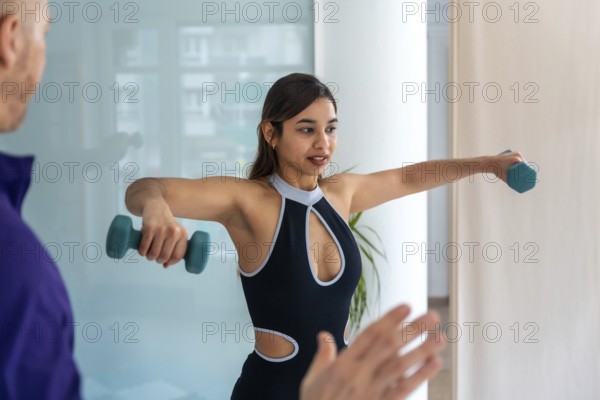 Young woman doing lateral dumbbell raises with a physiotherapist in clinic, focused on shoulder