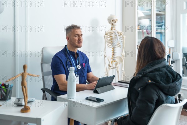 Male doctor in scrubs and stethoscope is talking with a female patient seated across his desk,