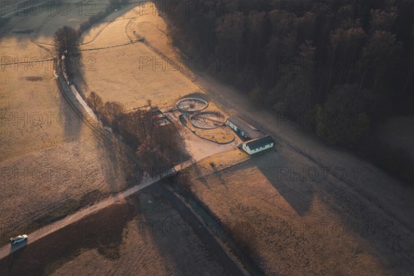 A path leads to a treatment plant with round structures, surrounded by fields and forest at sunset,
