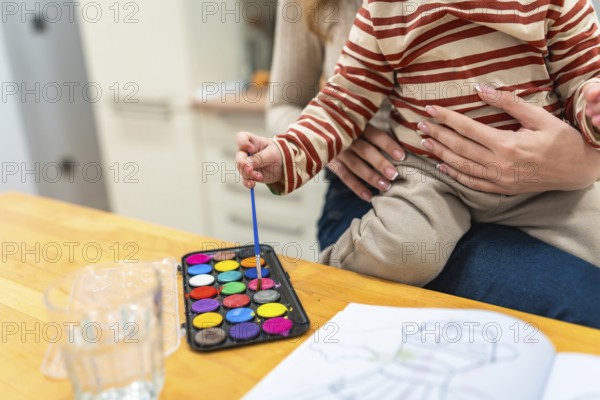 Young child held by mother, dipping a brush into a vibrant watercolor palette while coloring in an