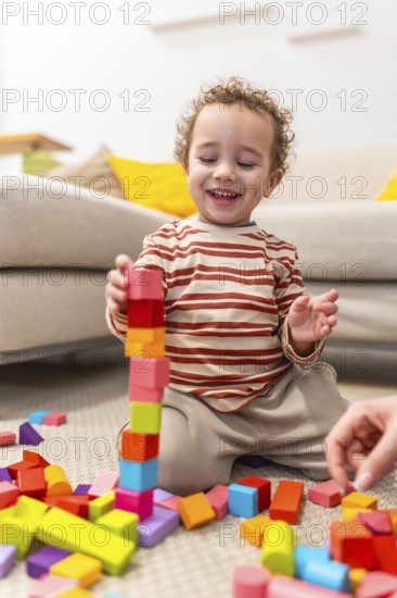 Happy toddler sits on carpet building a tall colorful wooden block tower, smiling and engaged in