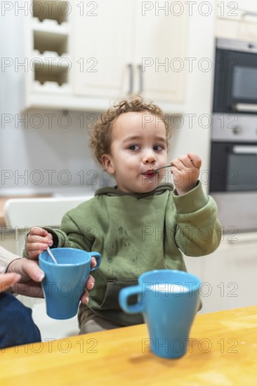 Curly haired toddler sitting at a kitchen table, independently eating yogurt from a spoon, leaving