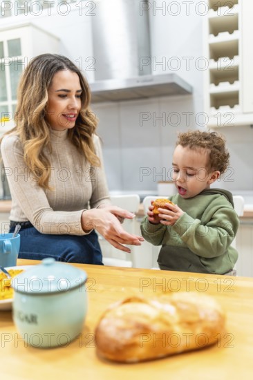 Mother looking at her happy child holding a muffin while having breakfast in a home kitchen,