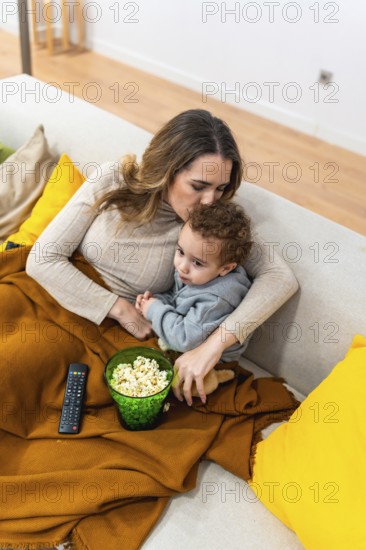 Mother and baby enjoying a bonding movie night at home, sitting comfortably on the sofa under a