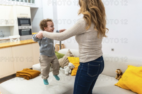 Happy mother holding her child's hands while they are jumping together on the sofa, enjoying a
