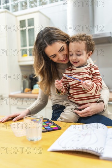 Mother holding her toddler son while they are painting with watercolors at a wooden table in a