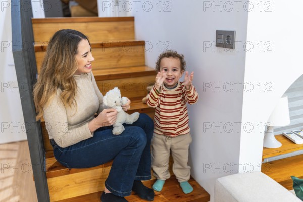 Mother and young son sit on wooden stairs at home, playing with a teddy bear and sharing a warm,