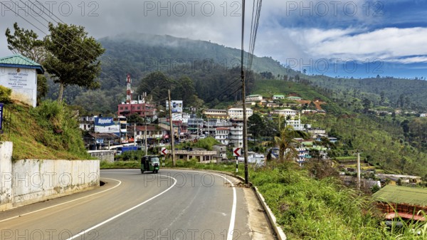 View of a curvy road in a village with hills and green countryside under a cloudy sky, The road on