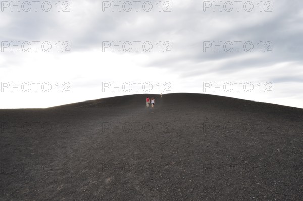 Wide landscape with people walking up a lava hill under cloudy sky, Craters of the Moon National