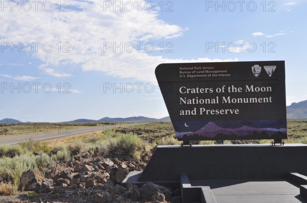 Craters of the Moon National Monument sign next to a road with wide landscape and blue sky, Craters