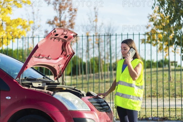 A woman wearing a reflective vest stands beside her car with an open hood, making a phone call