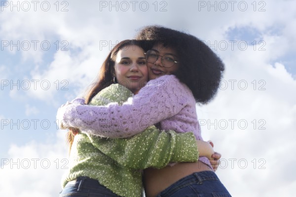 Two friends embrace warmly against a clear blue sky with fluffy clouds, wearing colorful knit