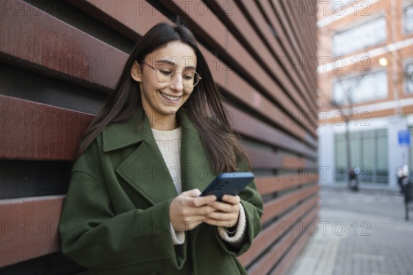 A woman wearing a stylish green coat leans against a wooden wall while engaging with her smartphone