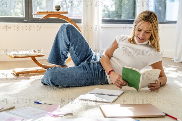 A woman enjoys reading a book while relaxing on a carpeted floor, surrounded by notebooks and a