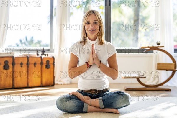 A woman dressed in casual attire, with a smile, meditates in a bright, sunlit room, positioned in
