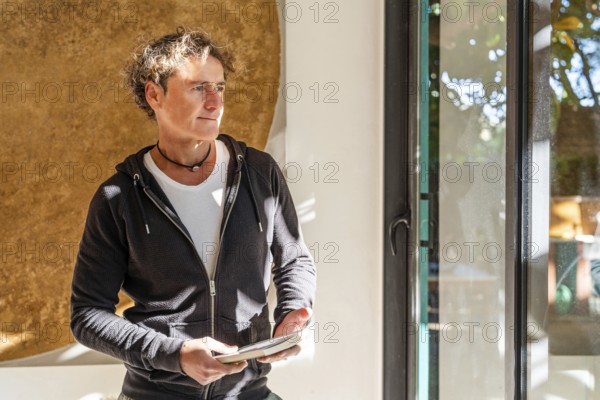 A relaxed man stands indoors holding a book, bathed in natural sunlight near a window Wearing a