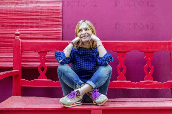 A cheerful young woman in a blue plaid shirt is seated on a bright red bench, smiling and looking