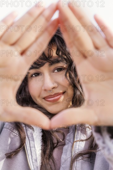 A young Latin woman with curly hair smiles through a hand frame, enjoying a sunny day Her casual