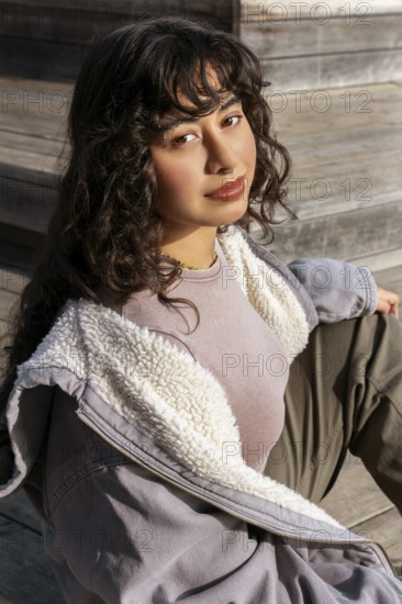 A young Latin woman with curly hair sits on wooden steps, enjoying a sunny day She wears a cozy