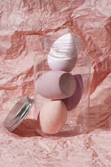Soft pastel cosmetic sponges in a transparent jar set against a crumpled pink paper background This