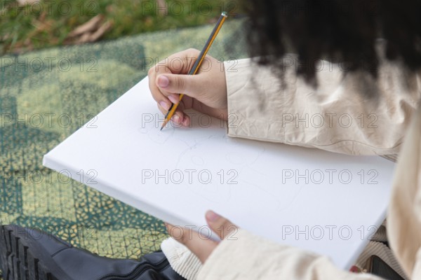 A young black woman sketches on a blank canvas using a pencil, surrounded by grass and natural