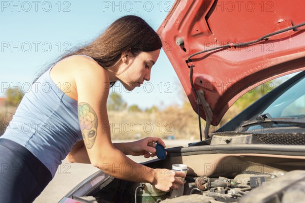 A woman in a blue tank top and tattoo on her arm examines her car's engine after a breakdown The