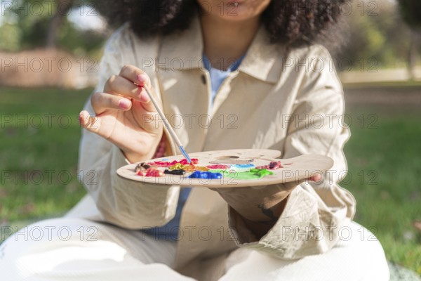 A young black artist holds a paintbrush and a palette filled with vibrant colors, engaging in plein