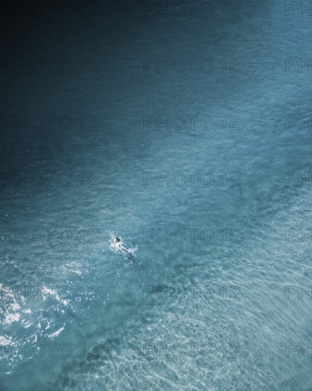 An aerial shot of a lone surfer paddling through calm turquoise waters along the Portuguese coast.