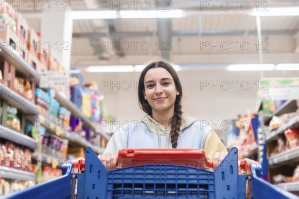A young woman looks directly at the camera while pushing a shopping cart in a supermarket aisle.