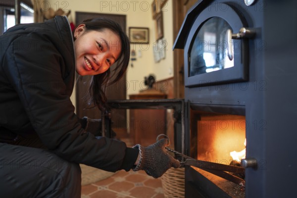 An Asian woman with visible joy looks directly at the camera while tending a wood burning stove