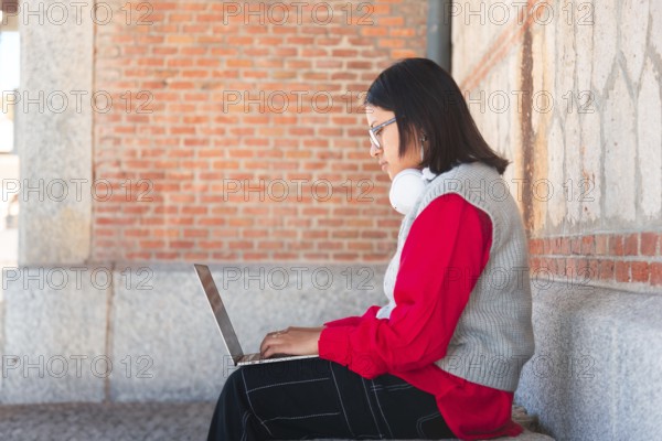 A Hispanic student with headphones focuses intently on her laptop while sitting against a brick