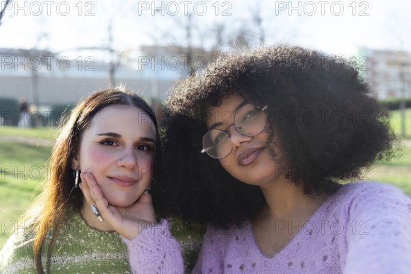 Two friends pose happily in a sunny park. One gently supports the other chin with a hand. They wear