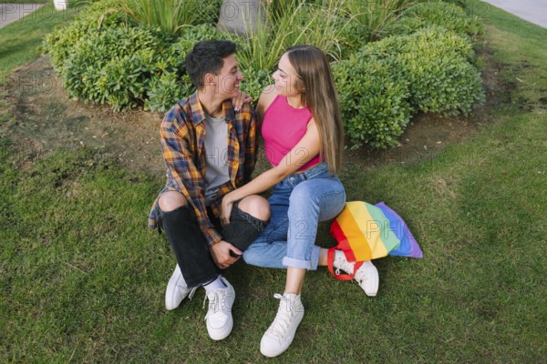 A joyful lesbian couple sits closely together on grass, embracing lovingly. A vibrant pride flag