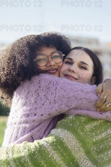 Two friends share a heartfelt hug, beaming with joy in a sunny outdoor setting. Both wear cozy