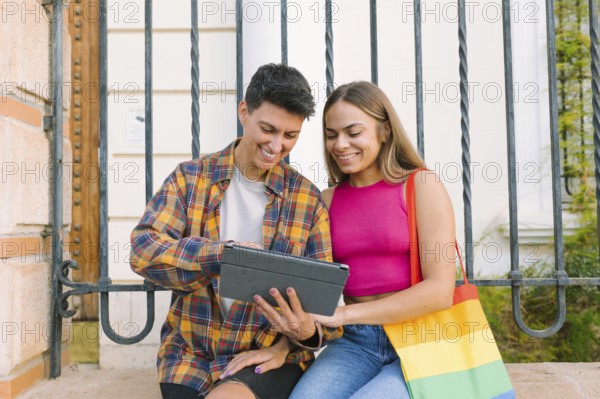 A happy lesbian couple sits together on a bench, smiling while looking at a tablet. One partner