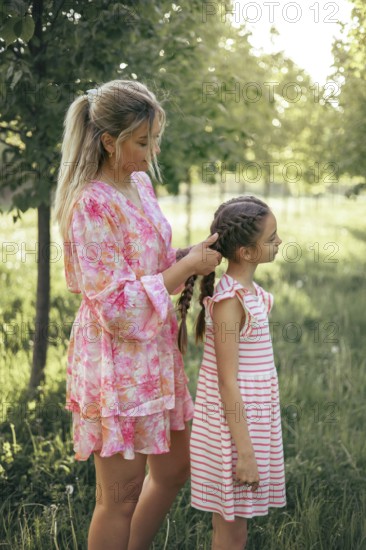 A loving mother gently braids her daughter's hair in a sunlit park. The tender moment captures