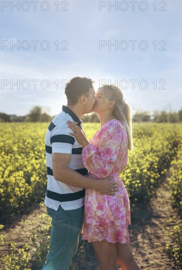 A loving couple embraces and shares a kiss in a golden flower field during sunset. The warm