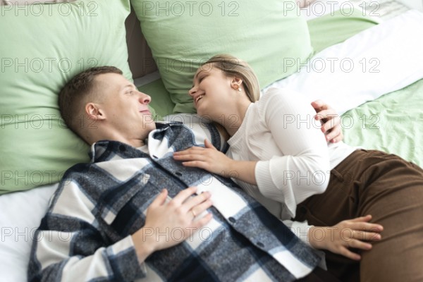 A young man and woman lie comfortably on a bed, sharing a close, intimate moment. They smile at