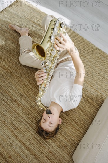 Young boy lying comfortably on a woven carpet and playing a saxophone. The cozy atmosphere