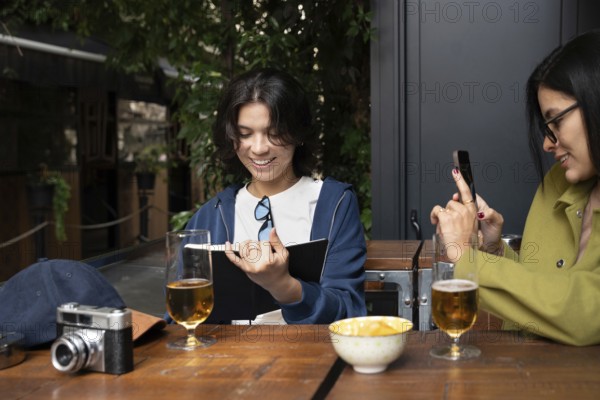 A lesbian couple shares a joyful moment at an outdoor cafe. One partner reads a book, and the other