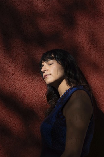 A Latin woman leans against a red wall with her eyes closed, basking in the sunlight. Shadows from
