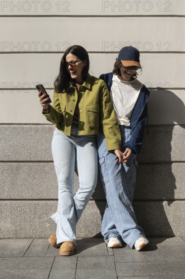 A joyful lesbian couple stands against a wall, holding hands and sharing laughter. The sunny day