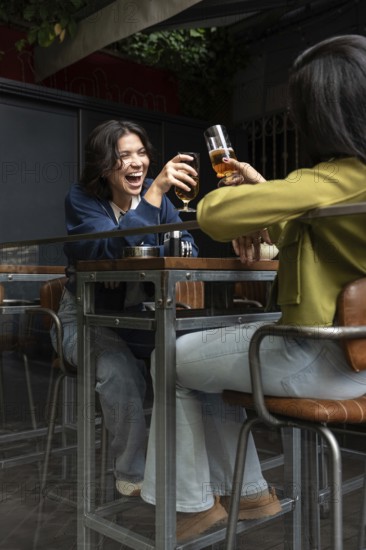 A lesbian couple joyfully toasts with glasses at a cozy cafe patio. They share a moment of laughter
