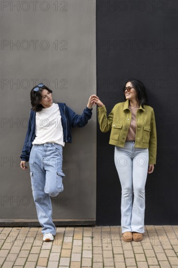 A joyful lesbian couple stands side by side, holding hands and smiling brightly while leaning