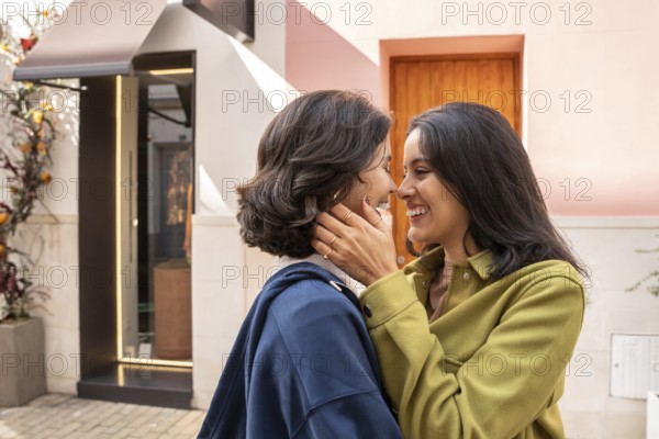 A lesbian couple smiling warmly as they embrace outdoors. The atmosphere is sunlit and joyful,